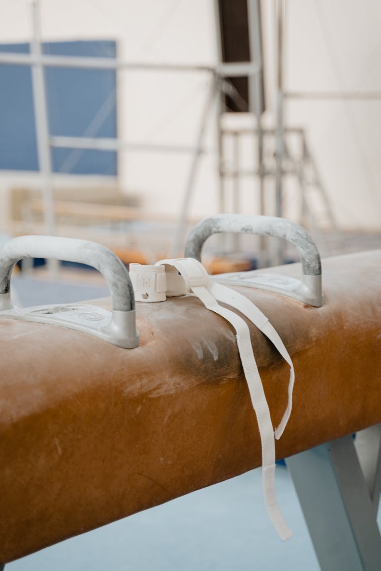 A detailed close-up image of a pommel horse with wrist wraps in an indoor gymnastics gym.
