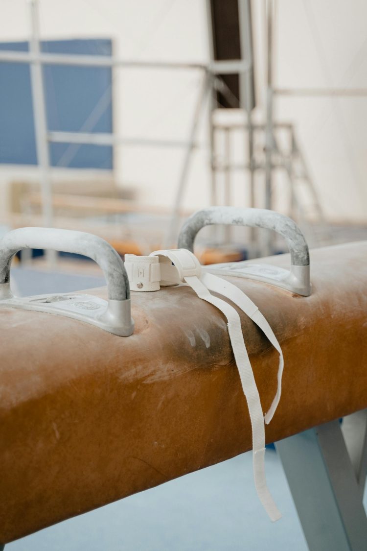 A detailed close-up image of a pommel horse with wrist wraps in an indoor gymnastics gym.