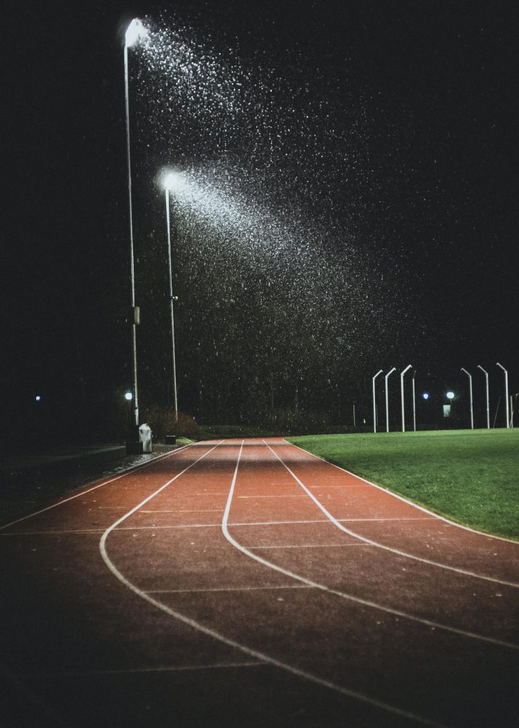 A track and field illuminated at night, with rain falling under the glow of streetlights.