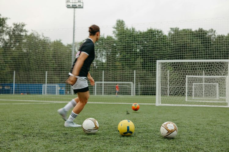 A woman practicing soccer on an outdoor field with multiple soccer balls and goals.